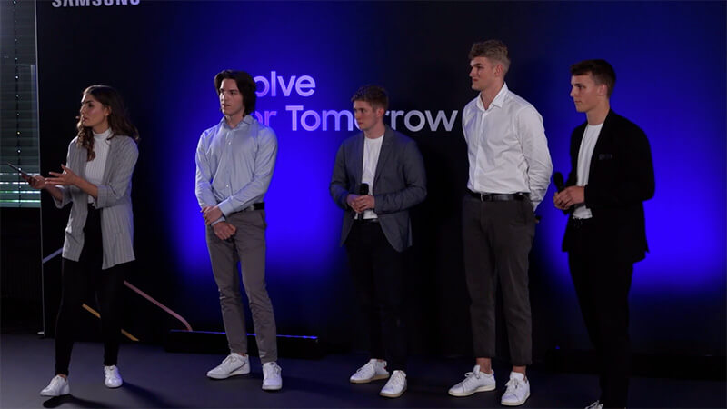 5 Moderatorin präsentiert auf Bühne vor vier jungen Männern bei Samsung Solve for Tomorrow Event mit blauem Hintergrund