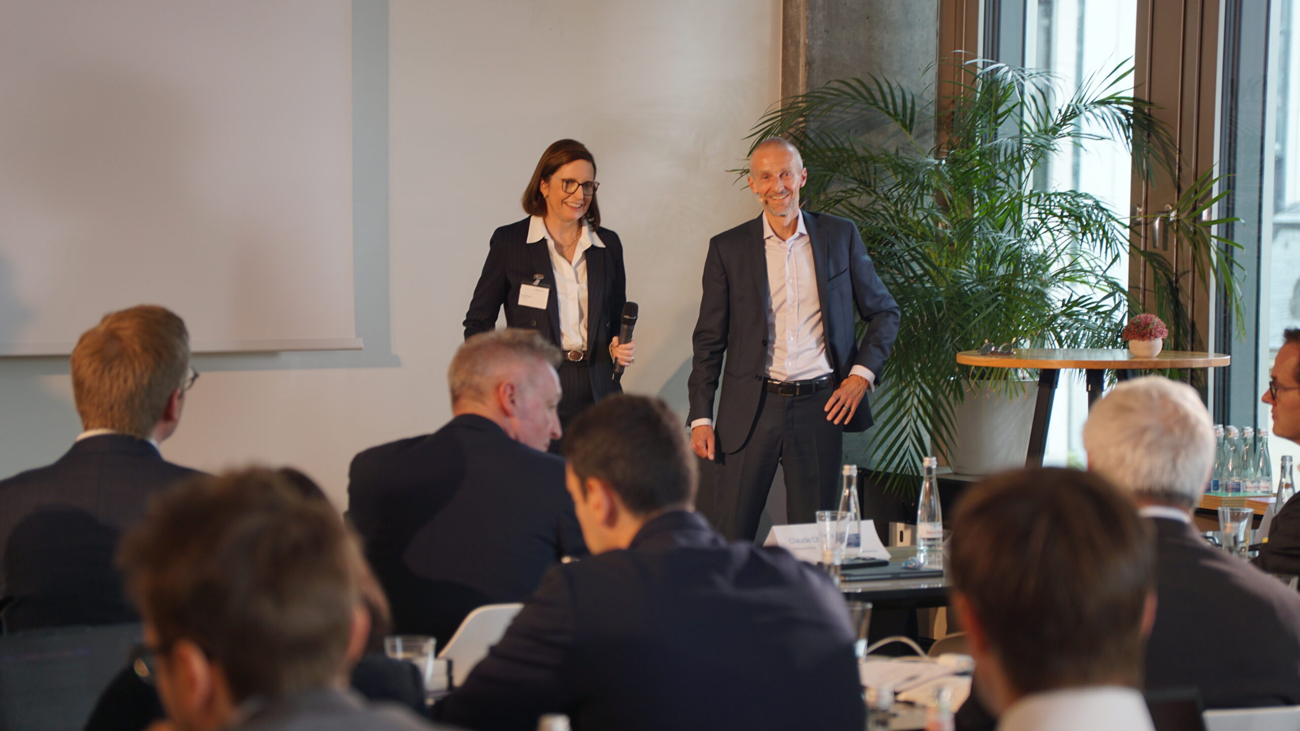 Moderatorin mit Mikrofon und Redner stehen vor Publikum bei Business-Veranstaltung in hellem Konferenzraum