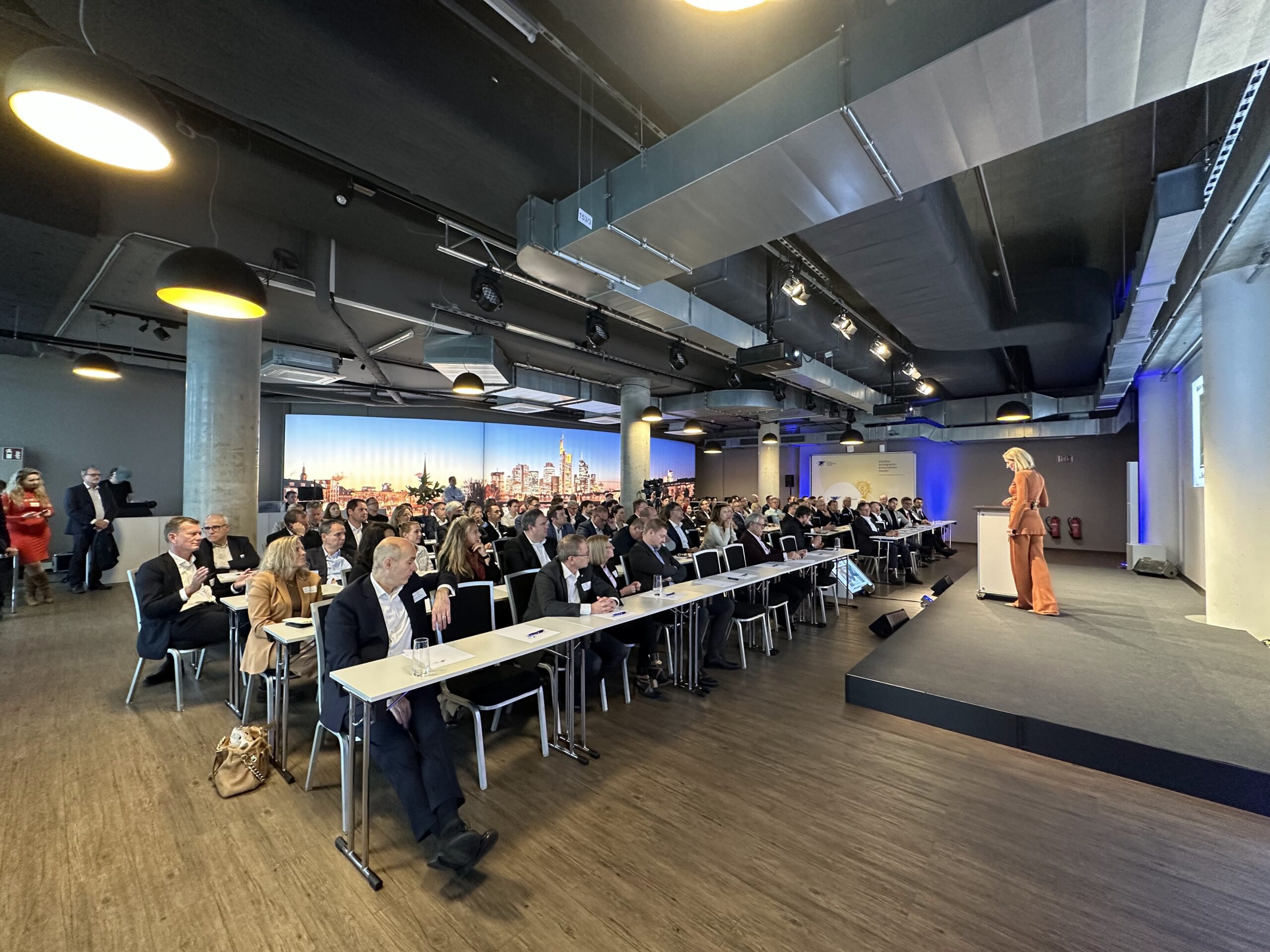 Rednerin in orangefarbenem Anzug präsentiert auf Bühne vor Publikum in modernem Konferenzsaal mit Skyline