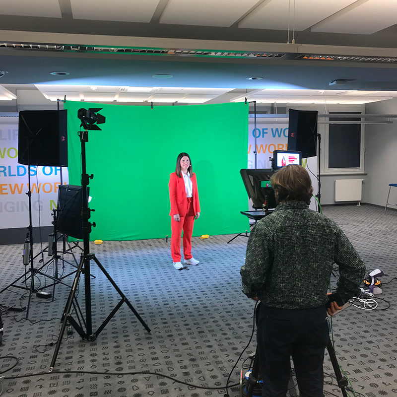 IMG_2003 Greenscreen-Videoproduktion: Frau in rotem Hosenanzug vor grünem Hintergrund, Kameramann im Vordergrund im Studio.