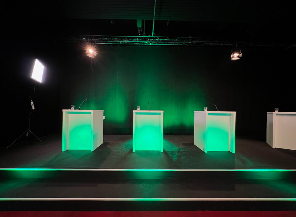 Bühnenaufbau mit vier weißen Pulten, grüner LED-Beleuchtung und professioneller Lichttechnik für Livestream im HV Studio2