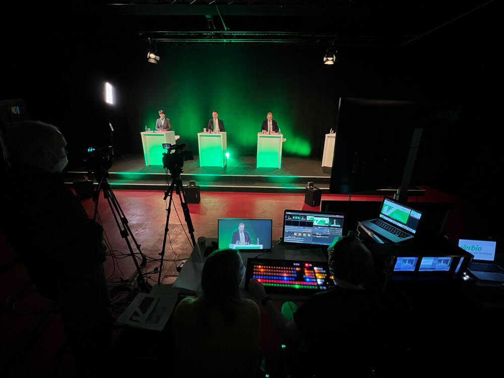 Blick in Verbio HV Studio 4: Bühne mit drei Pulten, grüner Beleuchtung und Streaming-Regie mit Monitoren und Kameras