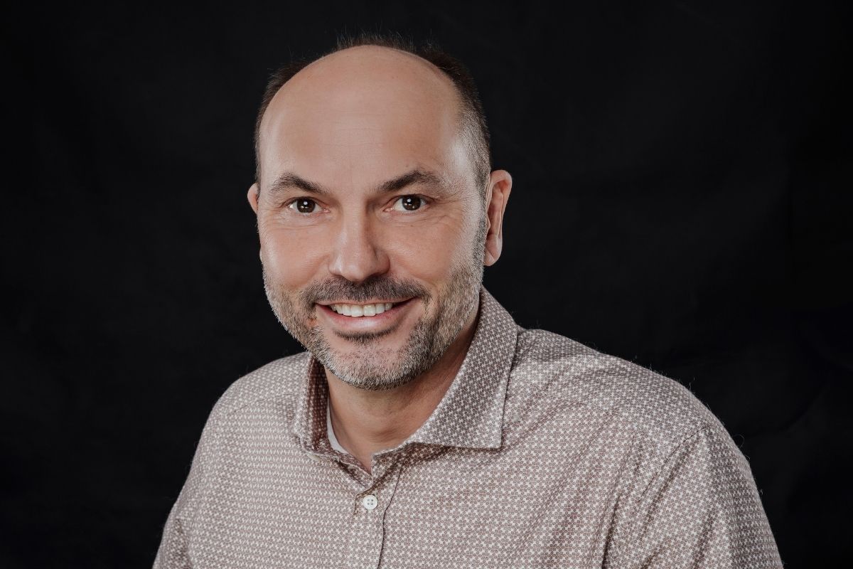 Porträt von Marc Ilgner, Gründer und CEO der NC3 GmbH, lächelnd vor schwarzem Hintergrund in braunem Hemd.