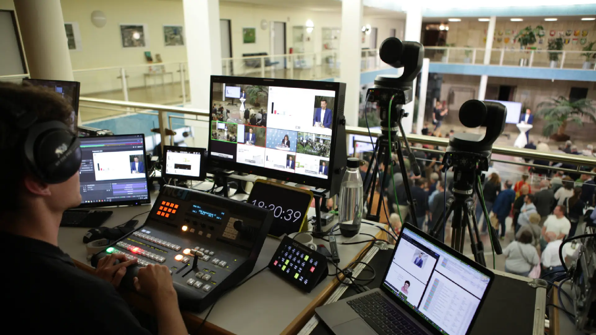 Techniker mit Headset bedient Mischpult in der Event-Regie, mehrere Monitore zeigen Live-Kamerafeeds, Kameras auf Stativen, Blick auf Bühne und Publikum in heller Halle.
