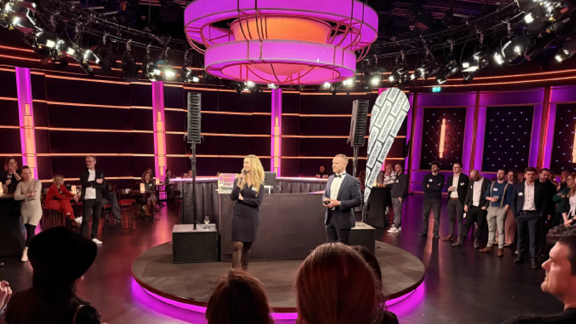 Moderatorin mit Mikrofon und Moderator im Anzug auf runder Bühne im NC3-Studio vor Publikum unter pinker Beleuchtung